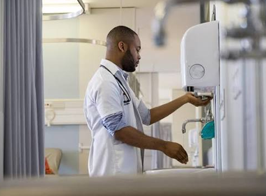 Healthcare professional wearing a white coat and stethoscope uses a wall-mounted water dispenser in a hospital room.